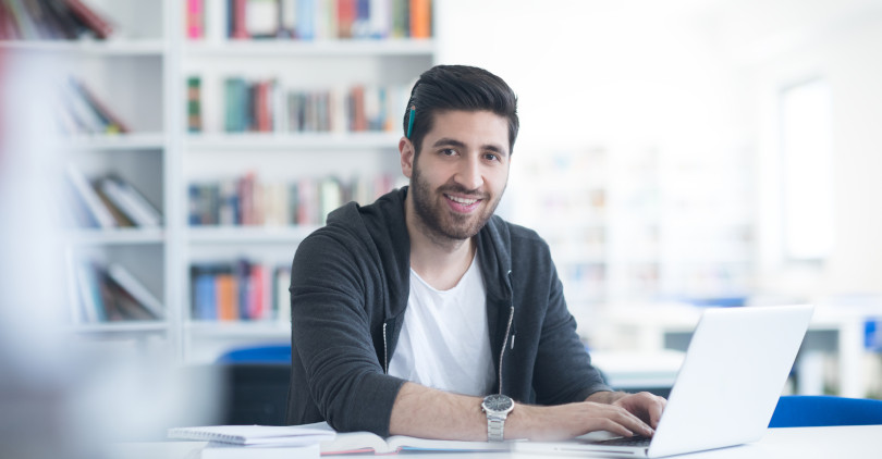 Empresario jovem com relógio e lápis na orelha sorrindo com notebook e estante de livros