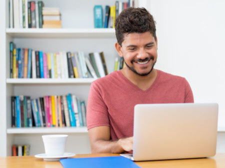 Homem de camiseta vermelha digitando no notebook e sorrindo com xícara de café ao lado e estante de livro ao fundo