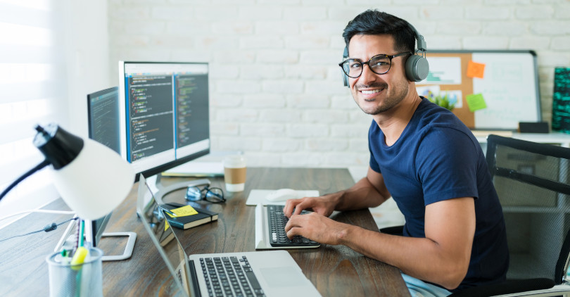 Homem jovem programador de óculos e camiseta azul escuro digitando com notebook no escritório com luminária