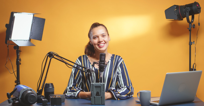 Mulher jovem blogueira infoprodutora de camisa listrada gravando e sorrindo com notebook xícara de café refletor de luz e microfone com fundo laranja