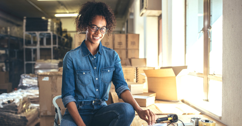 Empresária de ecommerce mulher jovem negra afro de camisa jeans azul sentada com caixas de estoque ao fundo