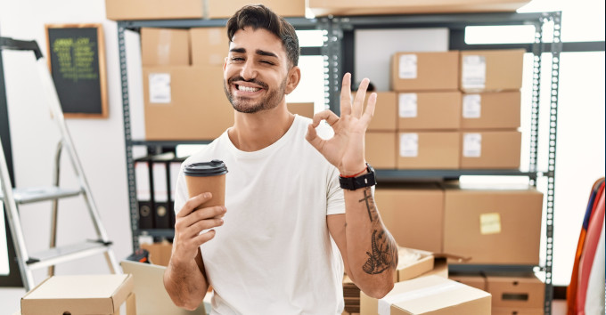 empresário jovem feliz sorrindo de camiseta branca fazendo gesto positivo e segurando café com caixas no estoque