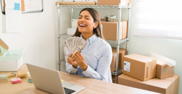 Mulher jovem empresária de ecommerce segurando dinheiro perto do notebook com caixas ao fundo